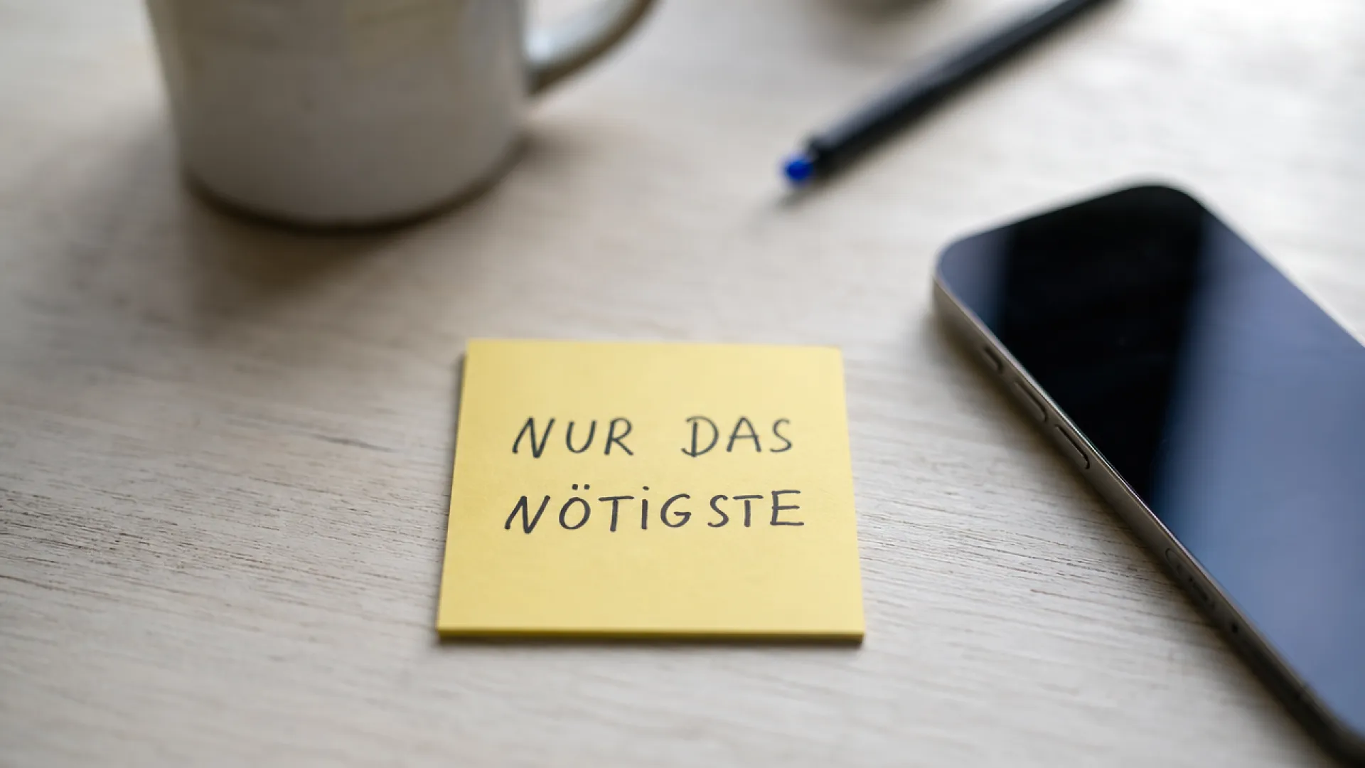 Ein gelbes Post-it mit dem Text Nur das Noetigste klebt neben einem Smartphone