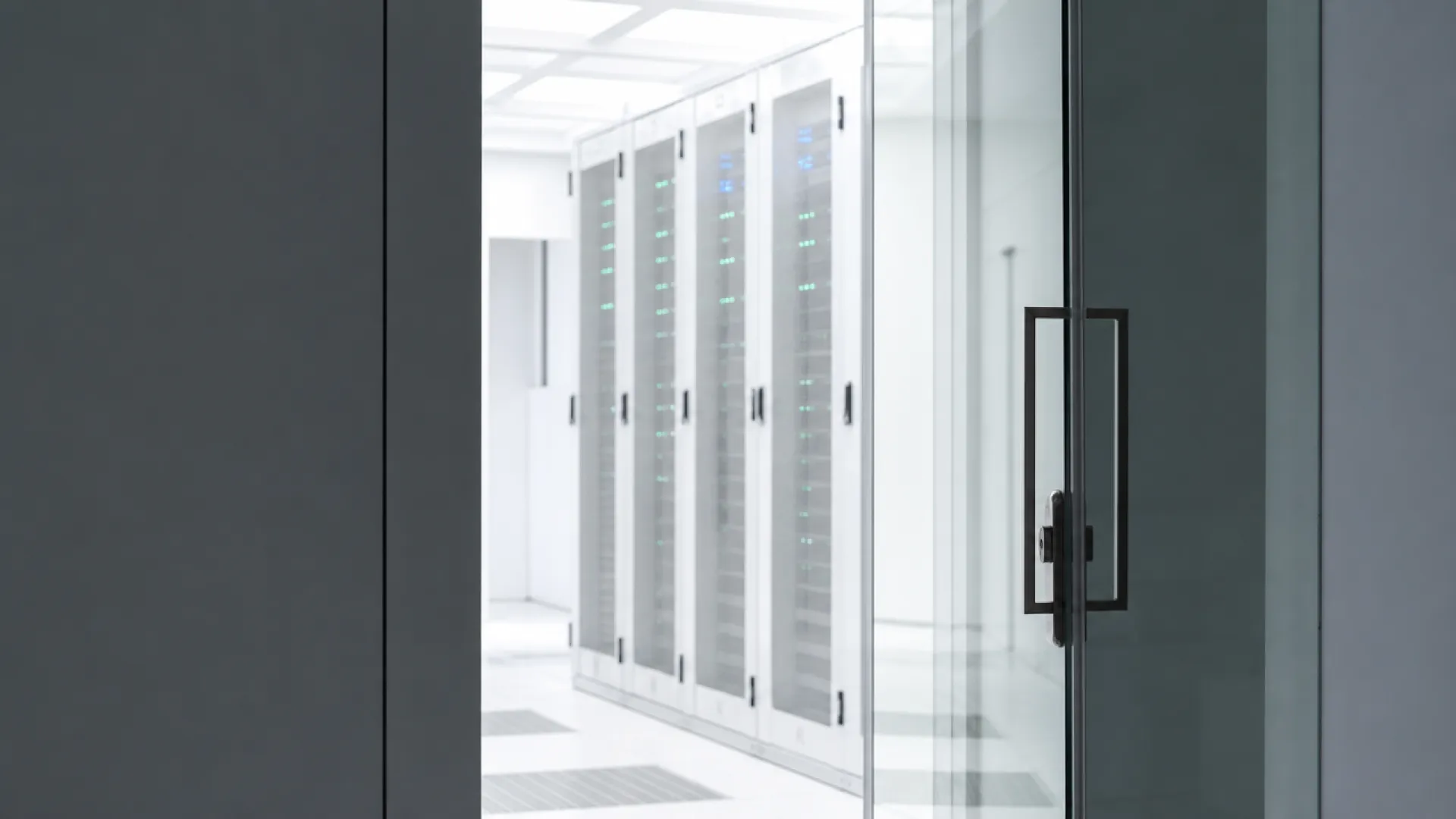 Blick durch einen Spalt einer dicken Glastuer in einen hellen Serverraum mit weissen Racks und leuchtenden blauen und gruenen LEDs